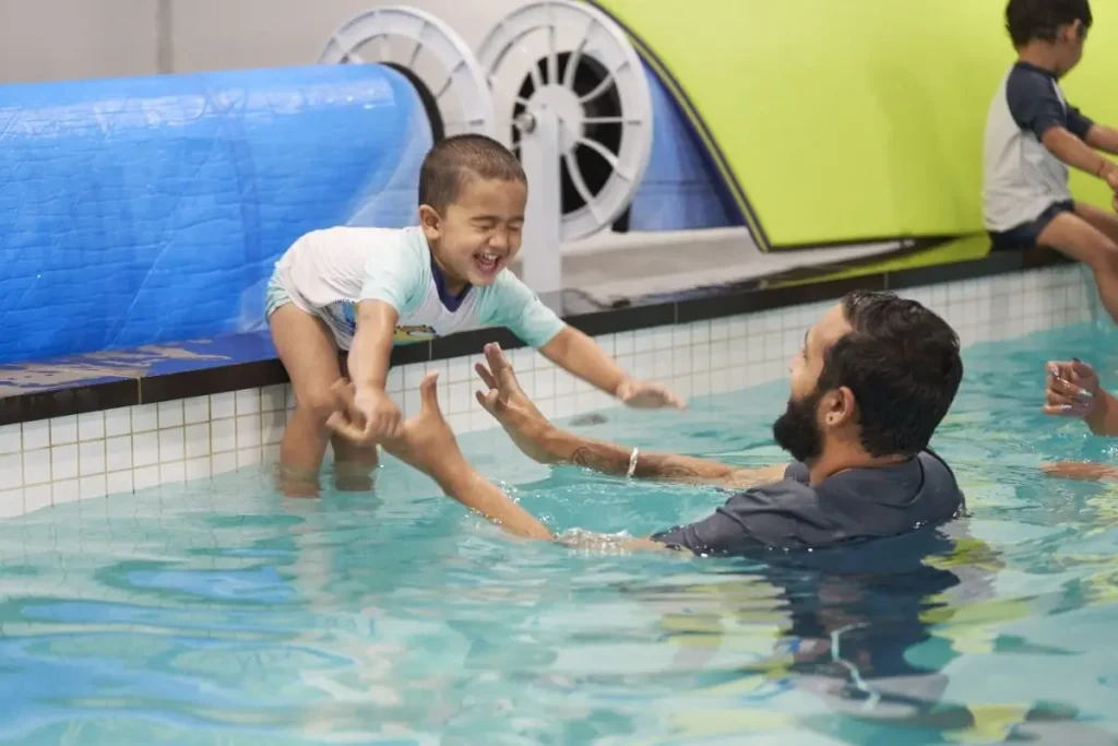 Young child smiling while practising a supervised pool entry during a swimming lesson, with an instructor ready to provide support and guidance in the water.