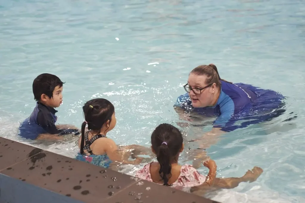 Swimming instructor leading a small group lesson with three young children seated at the pool edge, demonstrating skills and providing personalised guidance in the water.